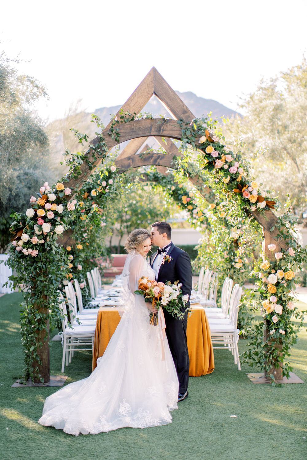 wooden arches decorated in lush florals with the groom kissing the bride's head underneath it all
