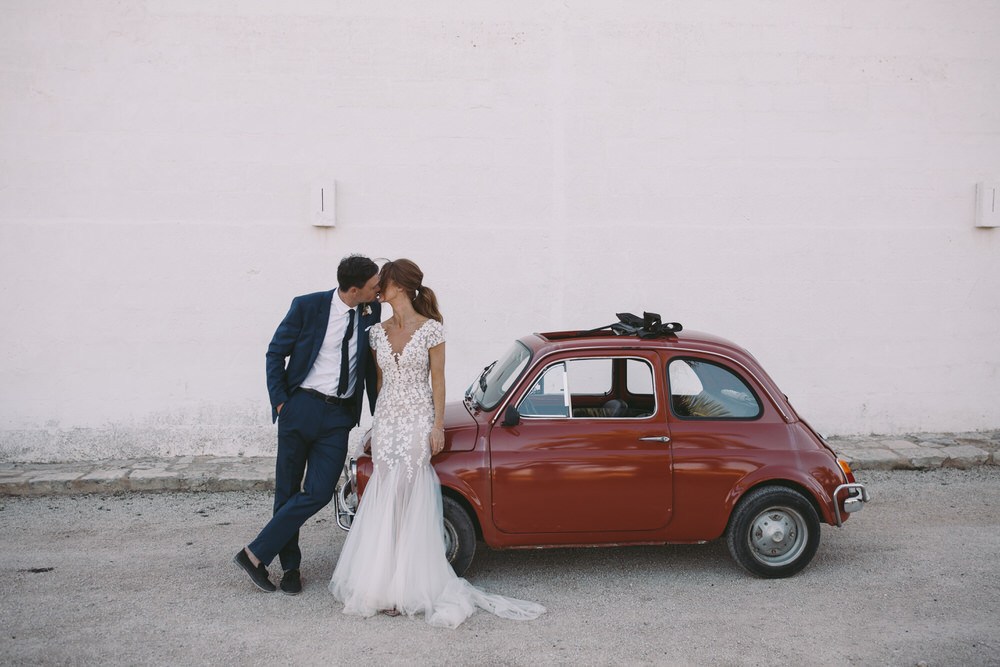 bride and groom kiss outside of their red getaway car